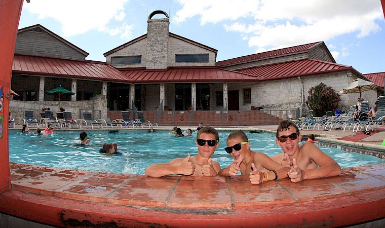 A sunny outdoor pool scene with swimmers lounging by the water and a red-roof building in the background.
