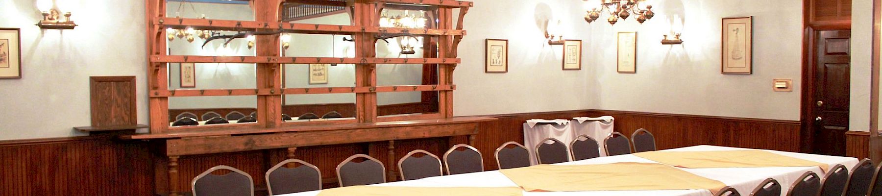 Bright dining room with white-clothed tables, dark chairs, ornate chandeliers, and a wooden bar or serving counter.