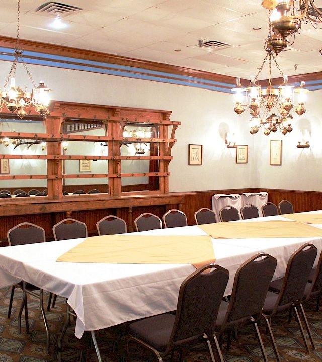 Bright dining room with white-clothed tables, dark chairs, ornate chandeliers, and a wooden bar or serving counter.