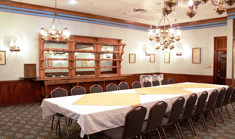 Bright dining room with white-clothed tables, dark chairs, ornate chandeliers, and a wooden bar or serving counter.