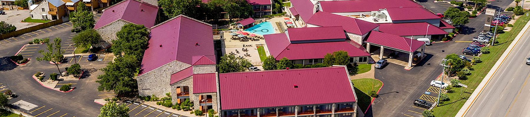 Aerial view of a suburban hotel complex with red roofs, parking lots, a pool, and tennis courts surrounded by greenery.