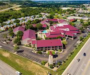 Aerial view of a suburban hotel complex with red roofs, parking lots, a pool, and tennis courts surrounded by greenery.