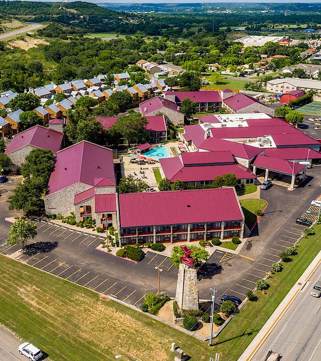 Aerial view of a suburban hotel complex with red roofs, parking lots, a pool, and tennis courts surrounded by greenery.