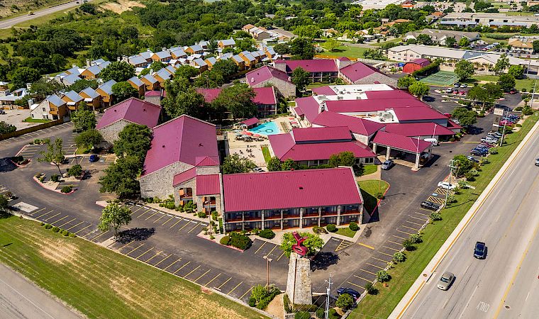 Aerial view of a suburban hotel complex with red roofs, parking lots, a pool, and tennis courts surrounded by greenery.