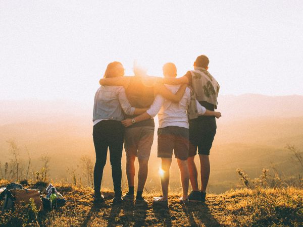 A group of friends stands in a sunny field at golden hour, hugging and facing the sunset with backpacks nearby.