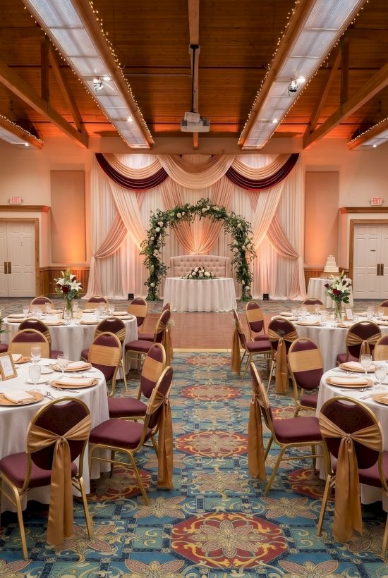 A wedding banquet hall setup with round tables, white florals, draped backdrop, and golden chairs under warm string lights.