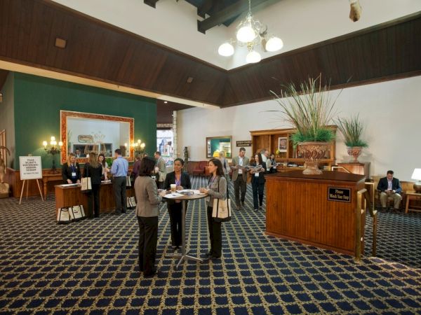 A hotel conference lobby with people networking, a front reception desk, and tall potted plants under chandeliers.