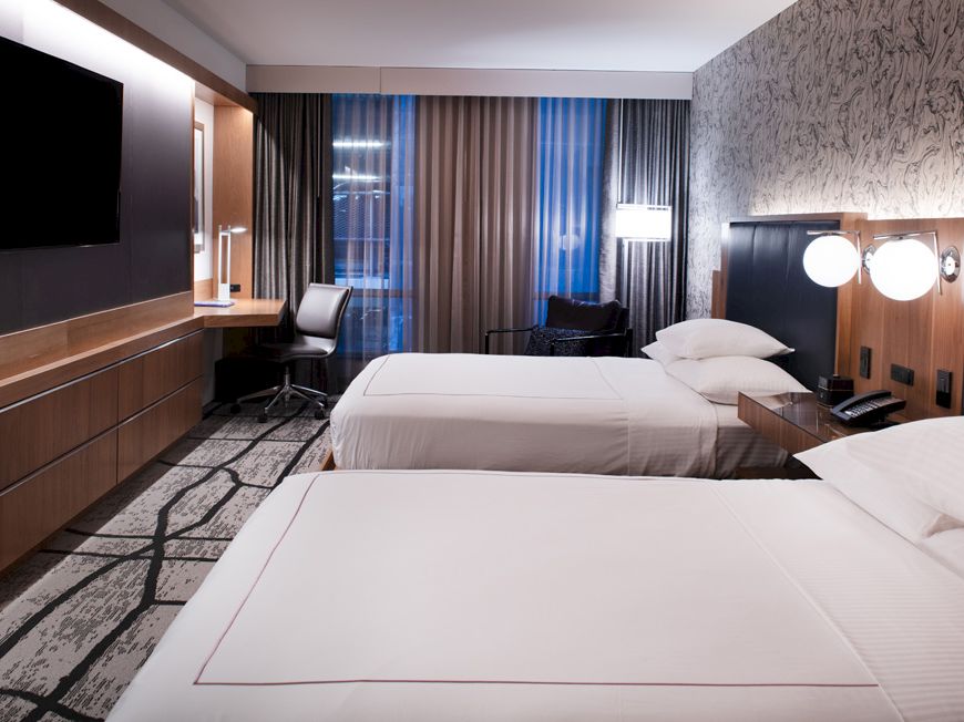 A modern hotel room with two white beds, a wall TV, desk seating, and dark curtains beside bright lamps.