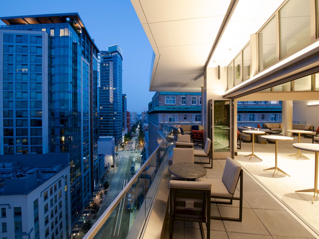 A high-rise rooftop lounge overlooks downtown, with patio chairs and tables beside a bright open terrace between skyscrapers.